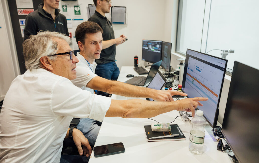 The image shows Marco Genovese (Head of Design) and Claudio Salomoni (Innovation Lab specialist) pointing to a monitor screen in Silverstone wind tunnel.