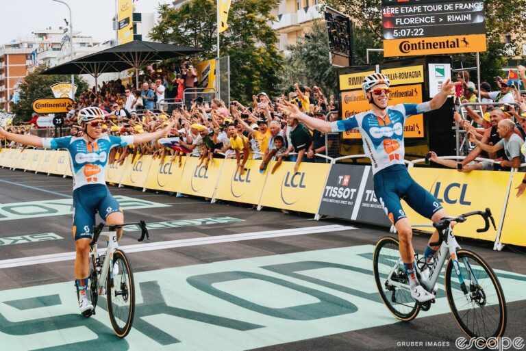 Frank van den Broek and Romain Bardet celebrate their 1-2 finish on stage 1 of the 2024 Tour de France. Both their arms are spread wide as they gesture to each other. Bardet is slightly ahead, and both have big smiles.