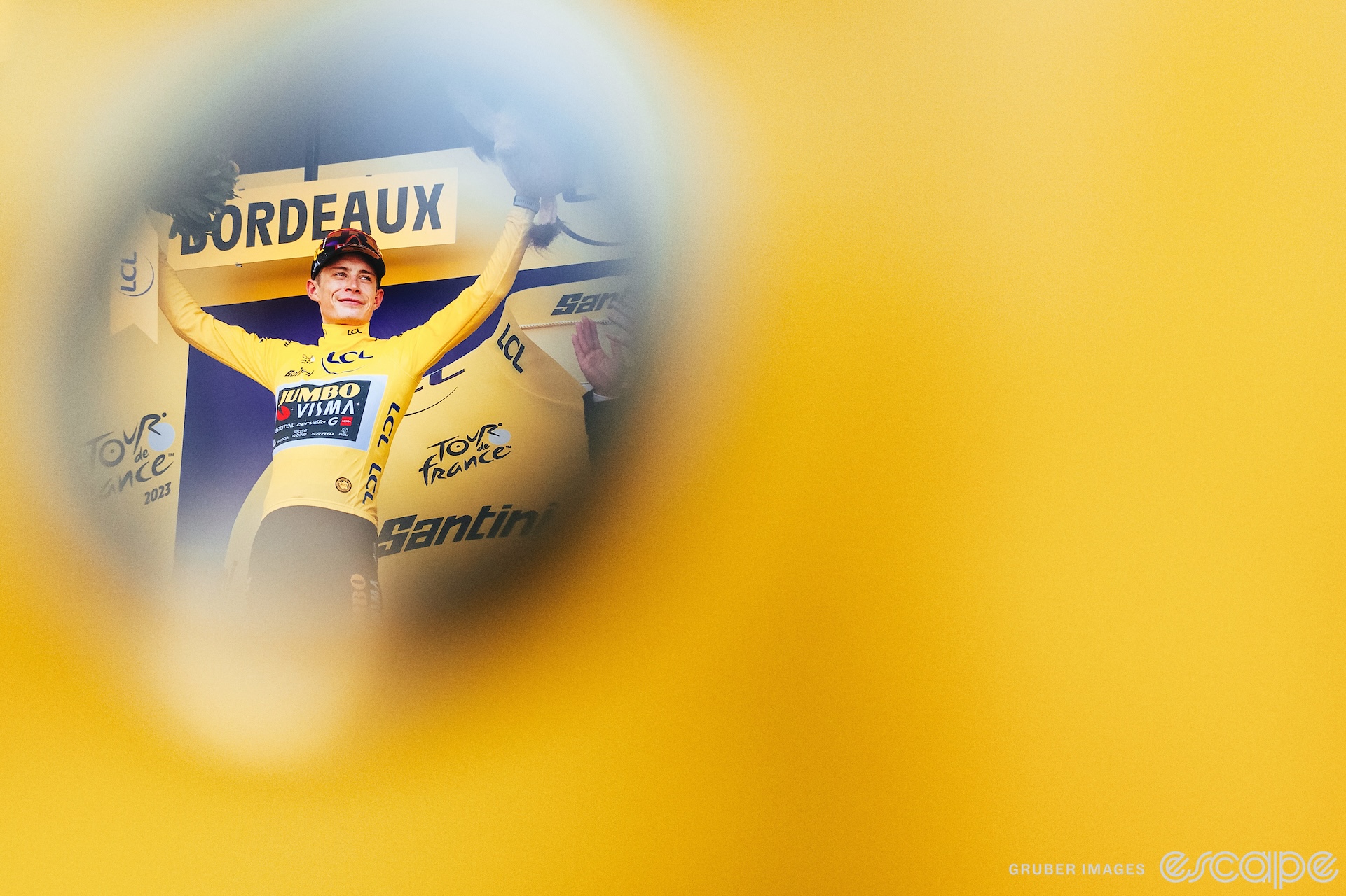 Jonas Vingegaard stands on the podium at the 2023 Tour de France in the yellow jersey with his arms raised. He's framed vignette-style through a hole in a blurry yellow flag or garment.