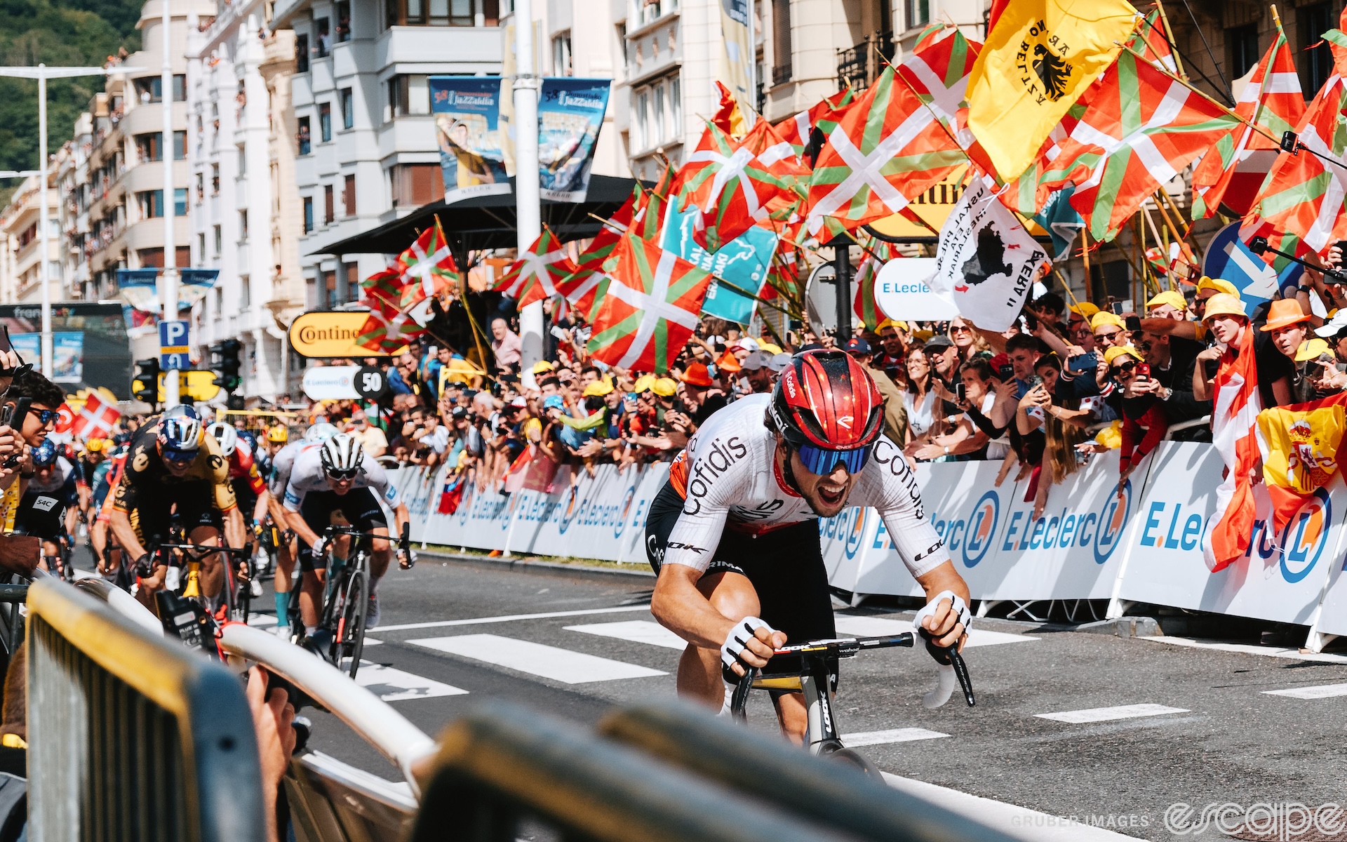 Victor Lafay sprints down the homestretch to win stage 2 of the 2023 Tour de France. His mouth is open and behind, Wout van Aert leads a furious chase. Masses of fans, many waving orange, green and white Basque flags, lean over the barriers to cheer.