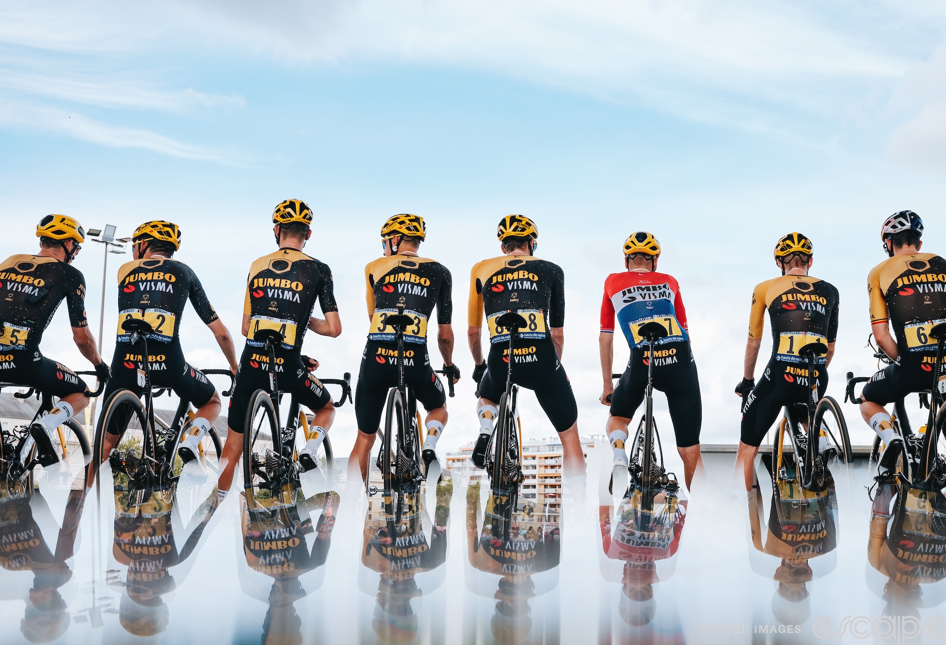 Jumbo-Visma stand on stage at sign-in during the Tour de France. The riders look out over an unseen crowd, and are shown reflected in the bottom third of the image in a trick of perspective