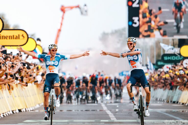 Frank van den Broek and Romain Bardet cross the line as Bardet wins stage 1 of the 2024 Tour de France. They're both smiling wide, shocked smiles and pointing at each other in pure joy. Behind, slightly blurred, a furious field finishes an unsuccessful chase.