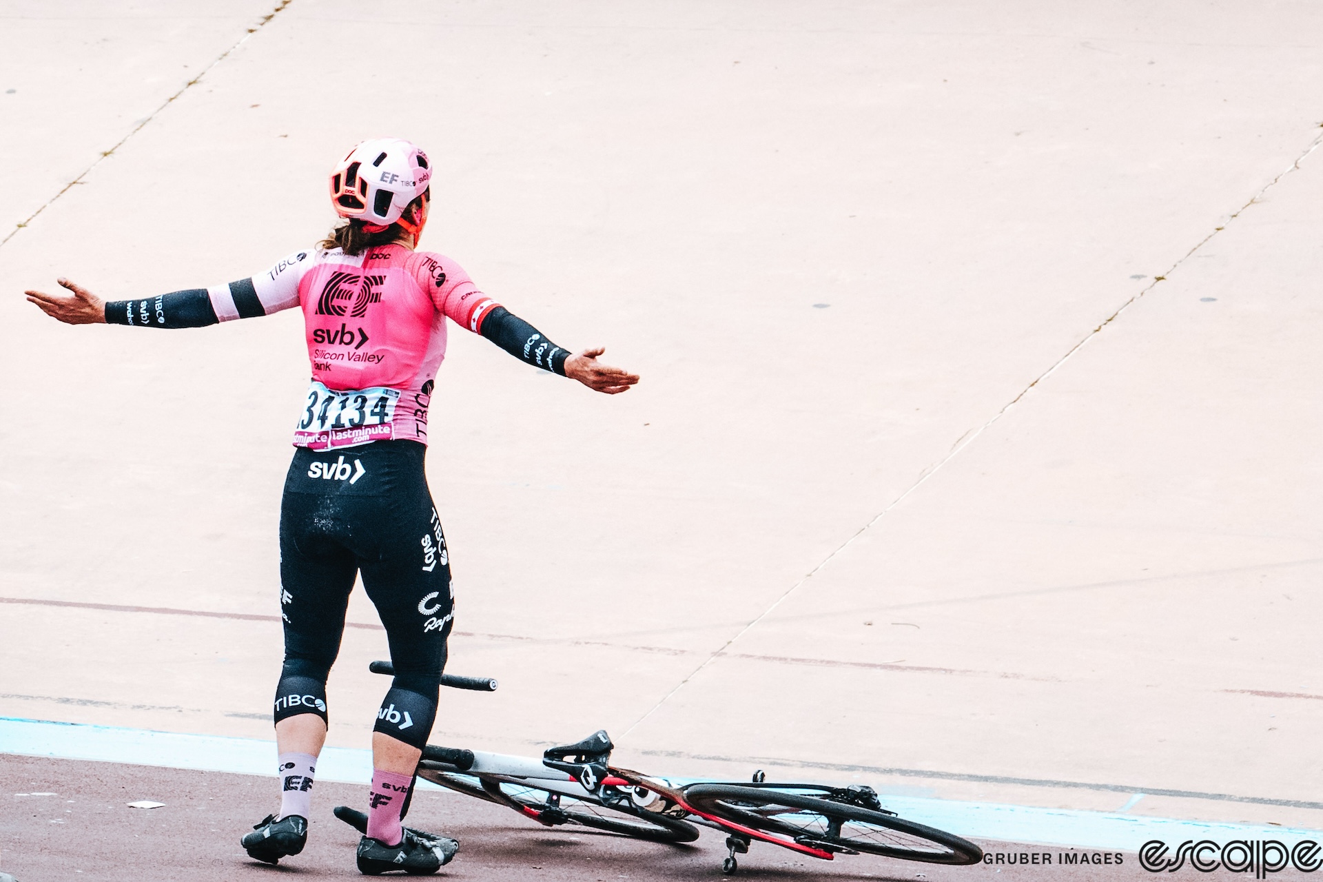 Alison Jackson throws her arms wide in celebration after winning the 2023 Paris-Roubaix Femmes. She stands in front of her bike, lying on its side in the velodrome, as she gestures to the crowd.