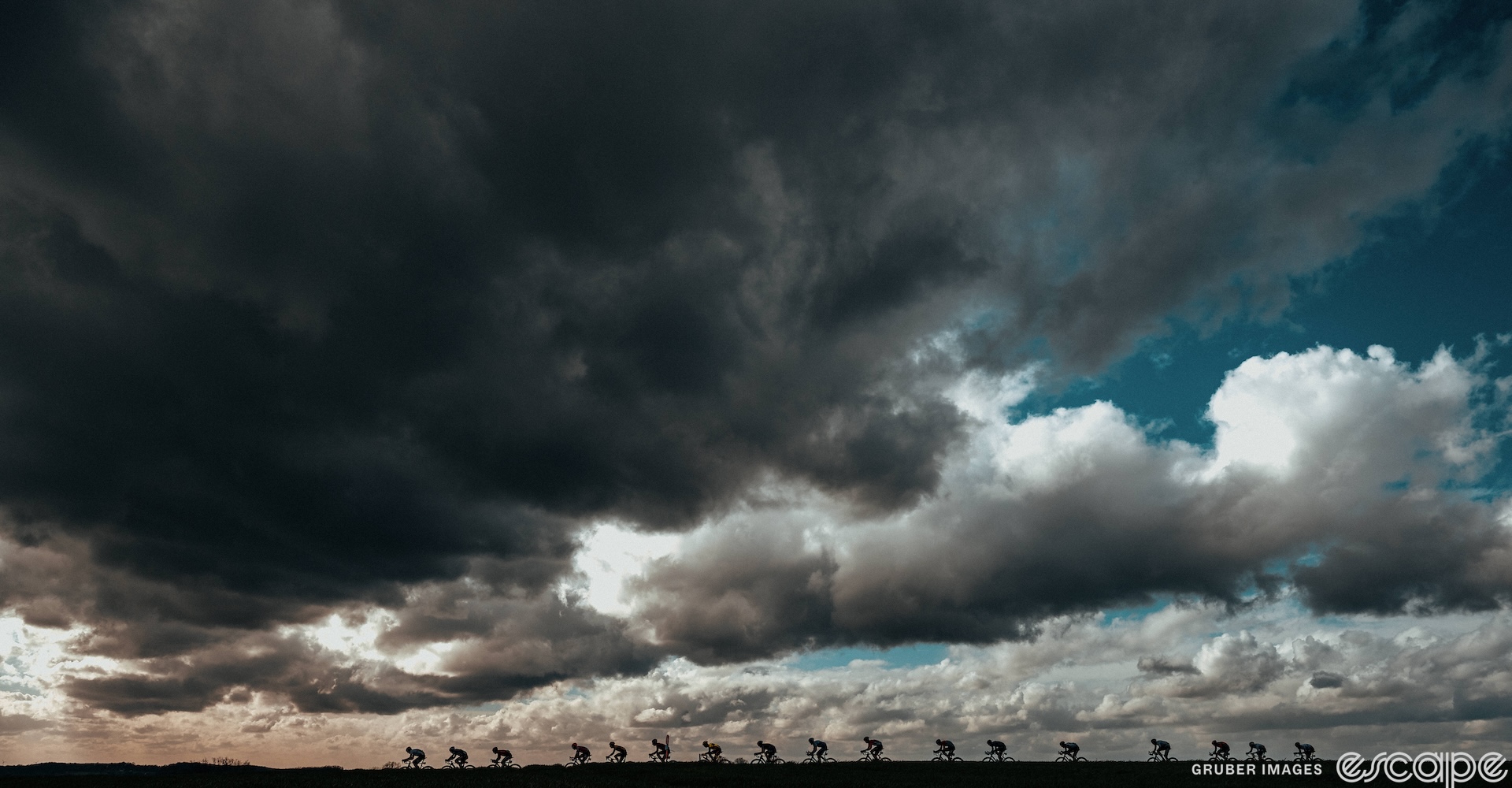 A line of racers stretches out in silhouette against a low, flat landscape. Dark, foreboding clouds loom overhead, broken by blue sky and streaks of sunlight. 