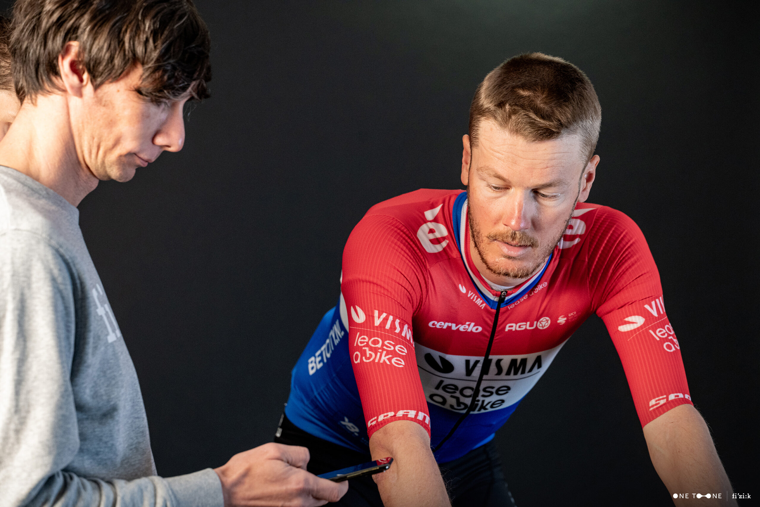 A bike fitter consults his phone to read pressure-mapping software as Visma-Lease a Bike pro Dylan van Baarle looks on while pedaling a bike during a fit session.