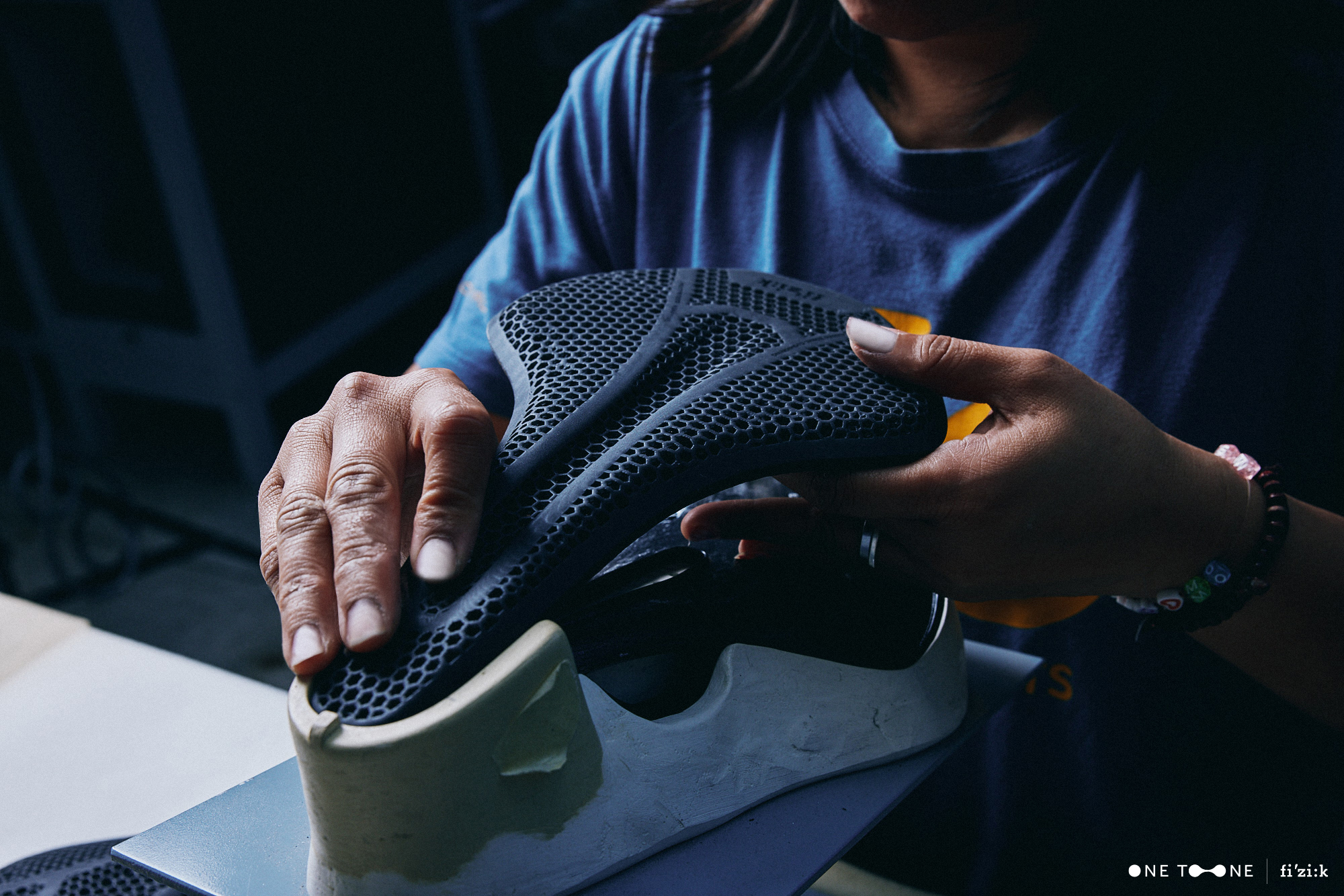 A worker's hands are shown as she fits a 3D-printed padding cover to a saddle shell at Fizik's HQ factory.