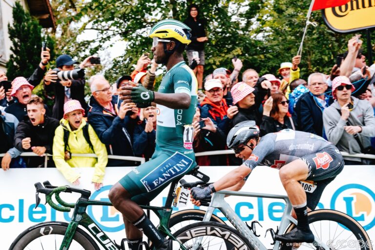 Biniam Girmay sits up as he wins stage 8 of the 2024 Tour de France. He's viewed in profile and to his right, Jasper Philipsen desperately throws his bike in an unsuccessful dash for the line.