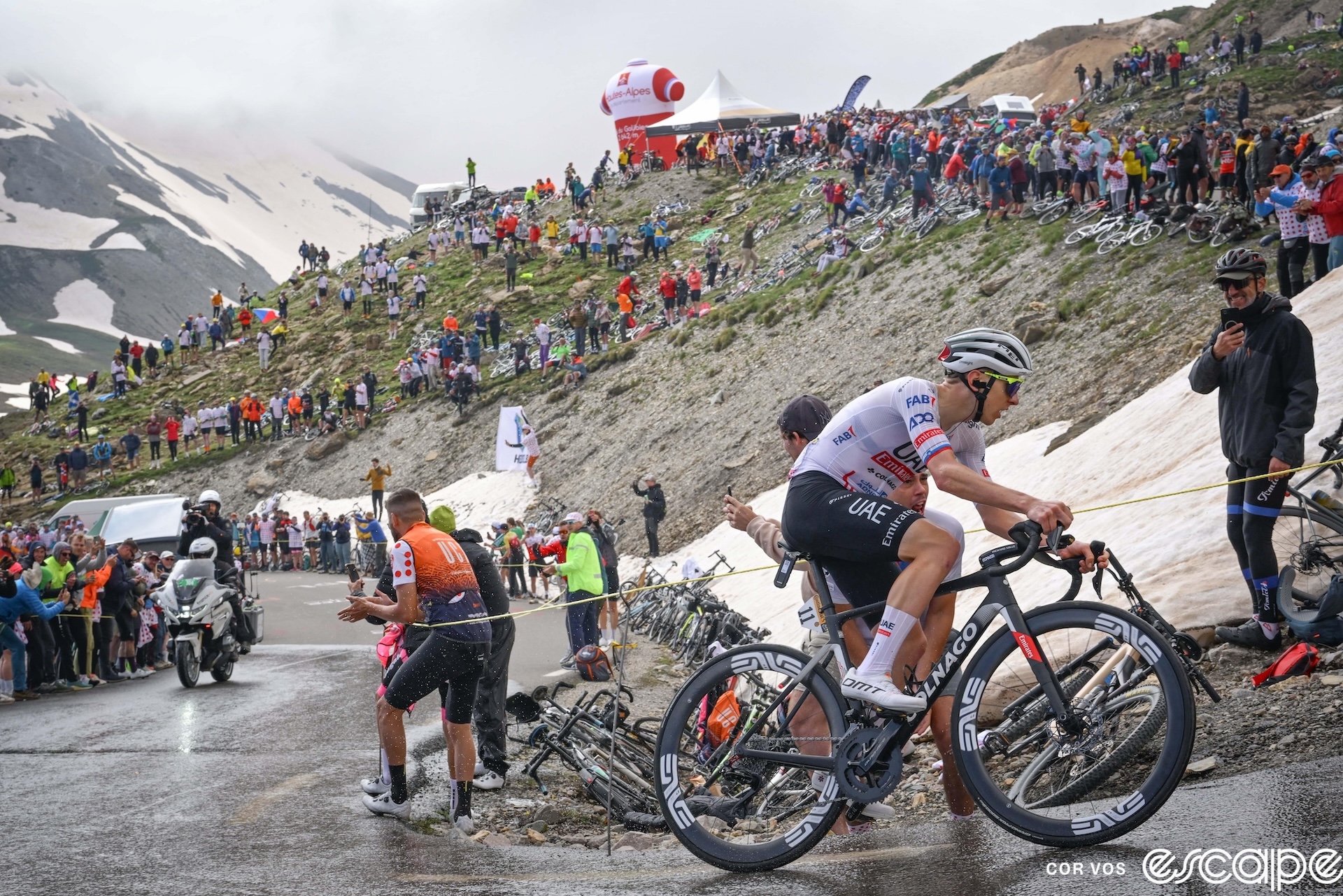 Tadej Pogačar rounds a wet switchback high on the Col du Galibier. The road is lined with fans and snowbanks and in the distance a snow-covered flank of a mountain looms.