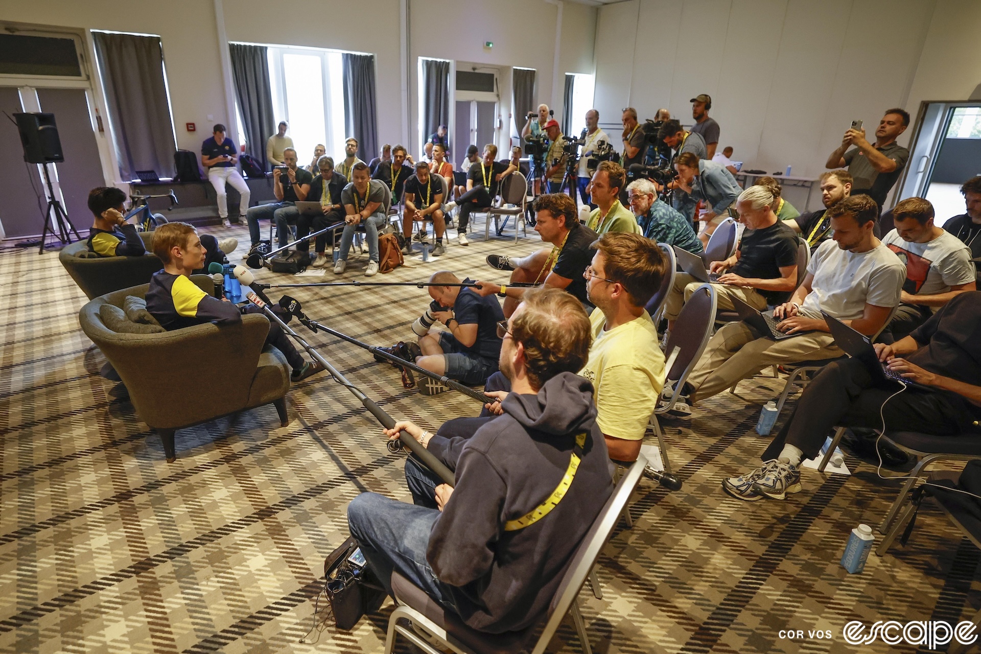 Wout van Aert and Jonas Vingegaard sit in large accent chairs in a conference room at a hotel. They're surrounded by a semi-circle of media, all pointing cameras and microphones as they answer press questions on a rest day.