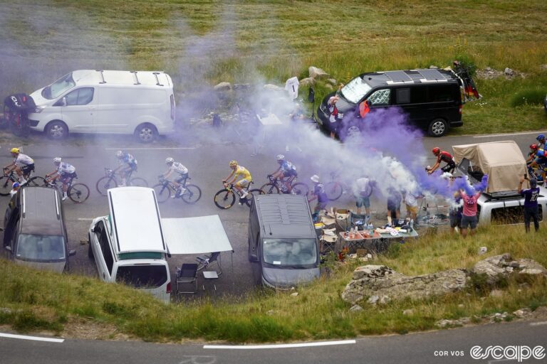 Fans line the road on stage 19 of the 2024 Tour de France. Seen from above, their camper vans are jammed into every available spot. Smoke from a grill rises near a table with a generous spread of food as Tadej Pogačar and other riders go past.