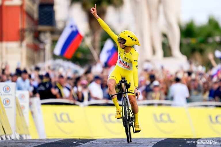Tadej Pogačar points to the sky in celebration as he rides to victory on stage 21 of the 2024 Tour de France, Slovenian flags waving in the background.