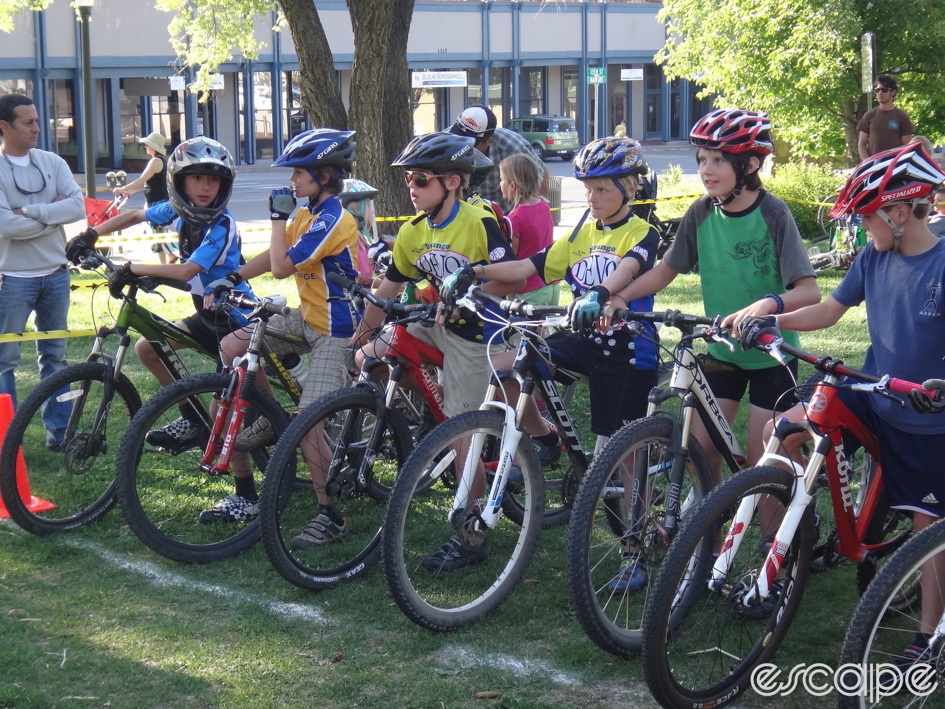 A group of kids lines up for a fun race in a Durango park. They're young, on green grass rather than a singletrack, and are dressed in a motley array of t-shirts and hand-me-down gear. One, far left, is in a full-face helmet.