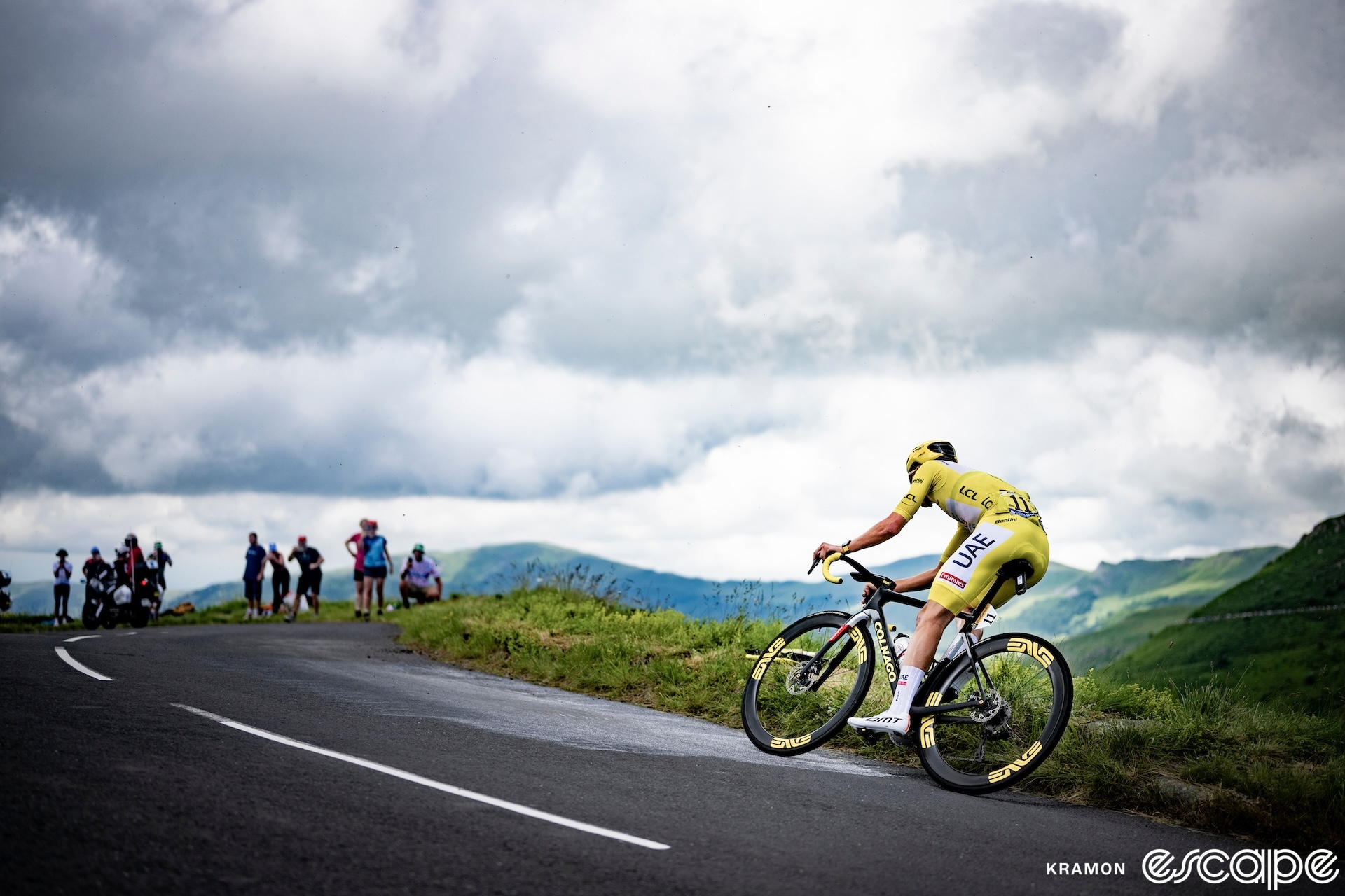 Tadej Pogačar rounds a corner on a slight uphill during a descent in the 2024 Tour de France. Clad in yellow, he leans into the turn as a group of fans await his approach.