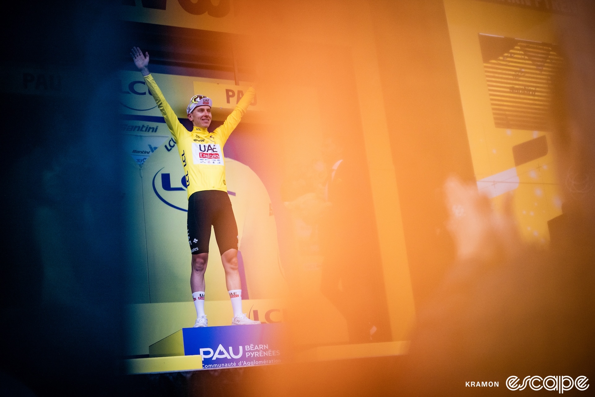 Tadej Pogačar stands on the podium of the Tour de France after a stage in Pau. He raises his arms in the yellow jersey and is seen framed through an orange haze created by the photographer.