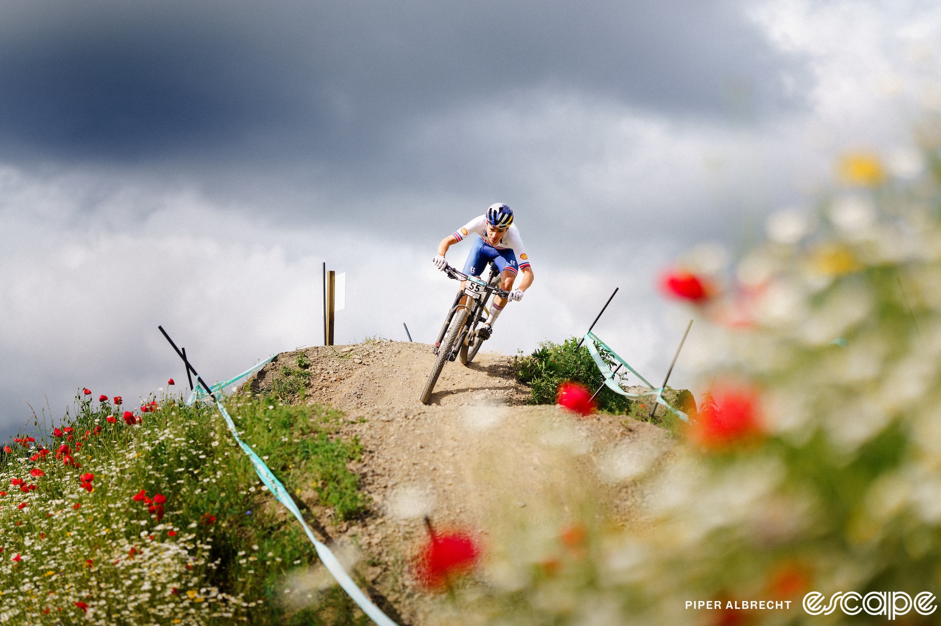 Tom Pidcock rides a berm on a descent at the 2023 World Championships in Glasgow, Scotland. He's framed by out-of-focus wildflowers, with dark skies behind.