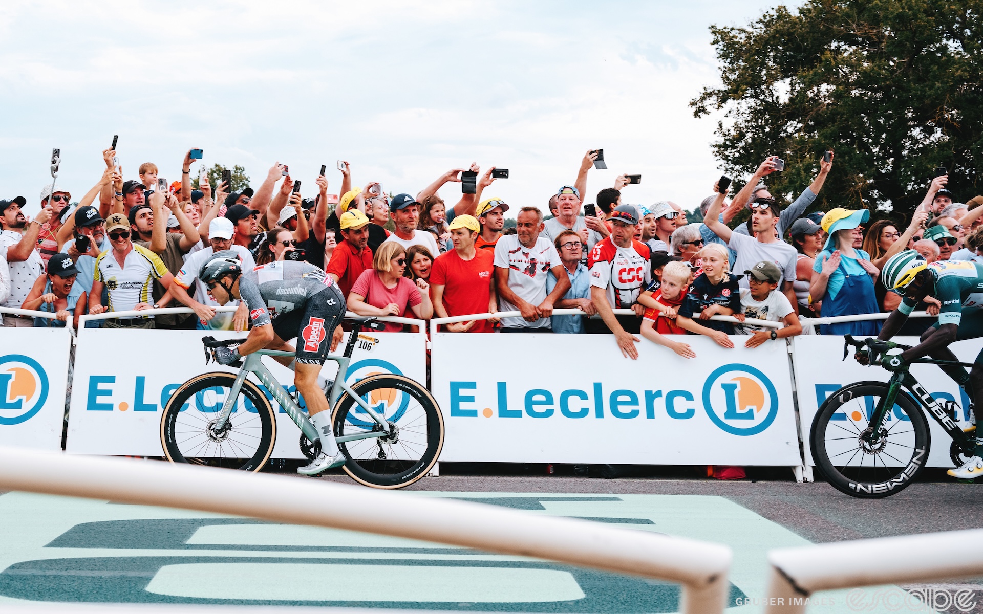 Jasper Philipsen is seen in profile sprinting for the finish line on stage 10 of the 2024 Tour de France. He's on the left side of the frame, fans leaning over white barriers with an "F Leclerc" logo. Biniam Girmay appears on the far right, emerging halfway into the frame in a futile chase. He is a bike length behind.