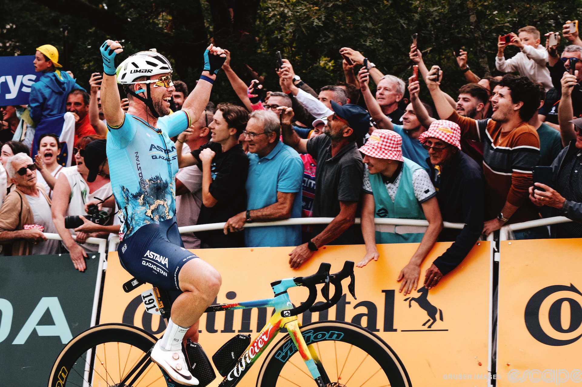 Mark Cavendish raises his hands as he crosses the finish line to win stage 5 of the 2024 Tour de France. He's shown alone, in profile, a wide smile breaking out on his face as fans slap the barriers and cheer.