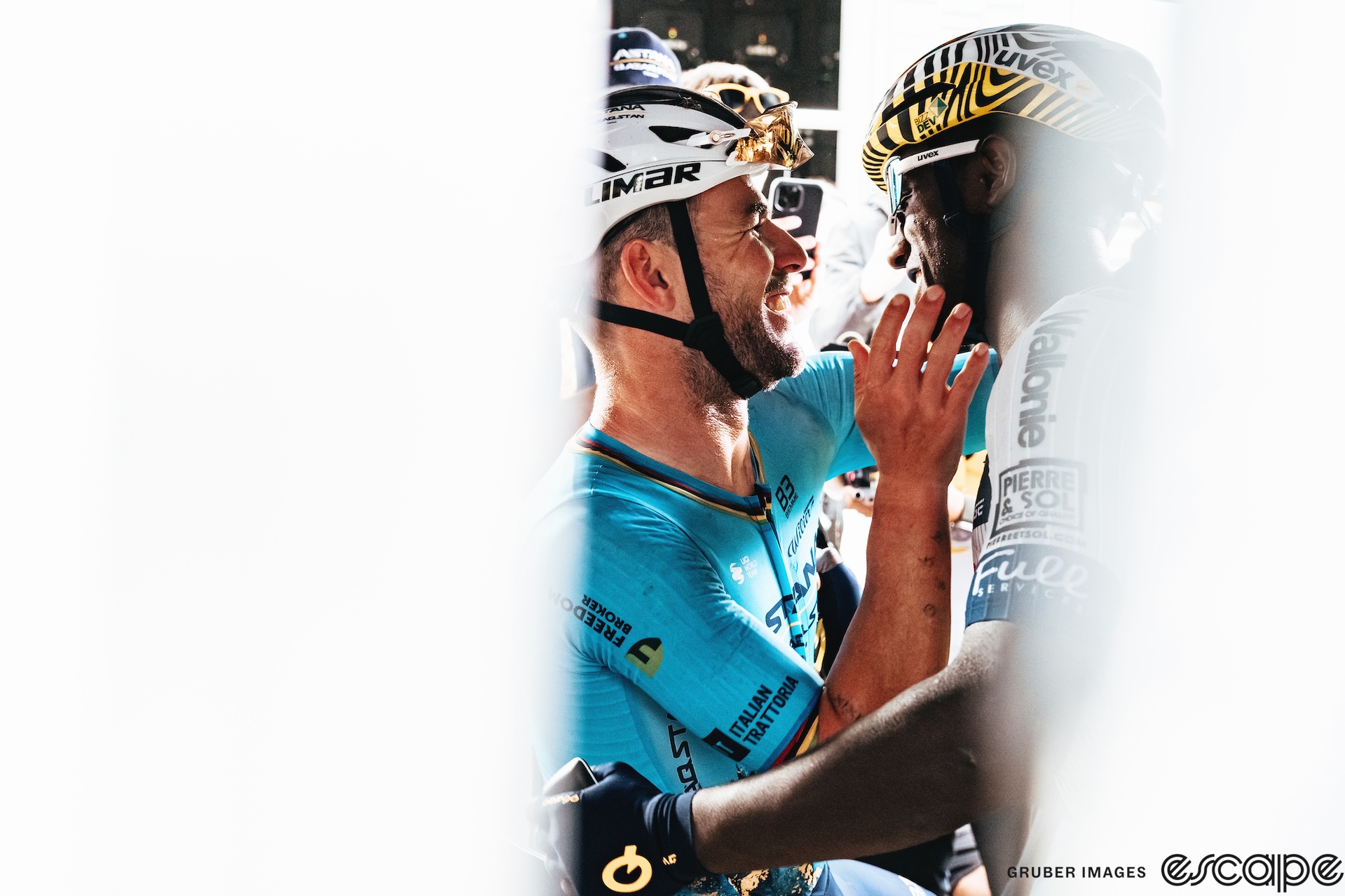 Biniam Girmay and Mark Cavendish share a momentary embrace at the finish after Cavendish's record-setting Tour de France stage win. Both are smiling big smiles, still with their helmets on, and are shown vignetted through white fencing that frames them in portrait profile.