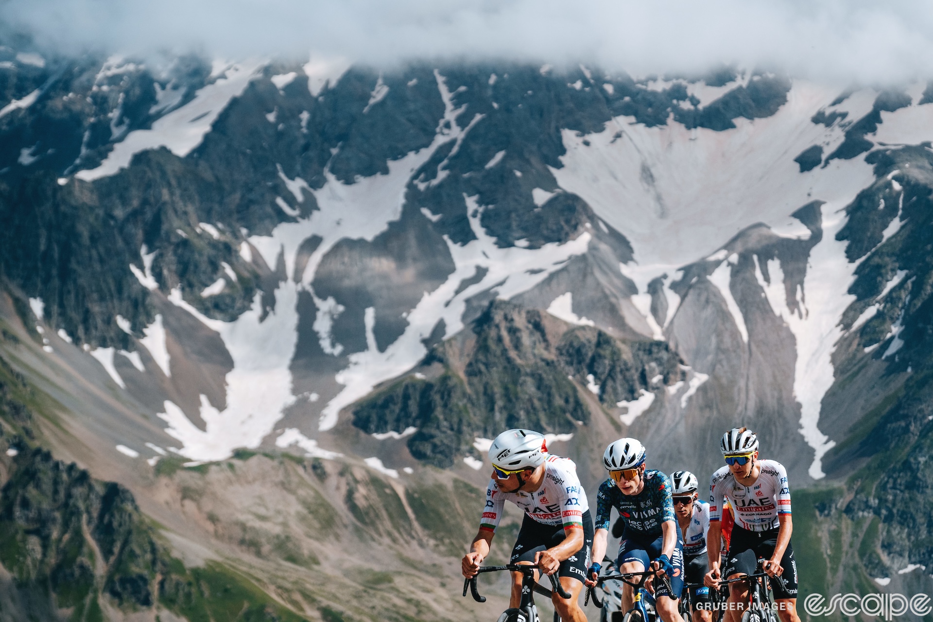 Joao Almeida leads Jonas Vingegaard, Tadej Pogačar, and Remco Evenepoel high on the Col du Galibier. The view down valley shows a sheer drop and a steep mountain filling the rest of the frame.