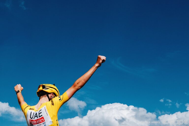 Tadej Pogačar raises his arms in victory as he wins a stage of the 2024 Tour de France. He is wearing the yellow jersey of race leader, with a matching yellow helmet. He is seen from slightly behind and below, framed against a blue sky with a lower border of clouds, as if to suggest that he is on another level from any other bike racer.