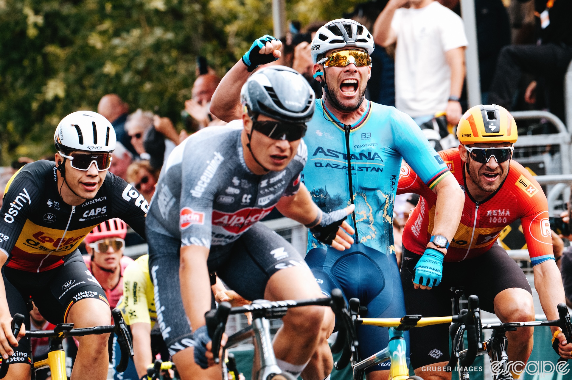 Jasper Philipsen reaches a hand back to congratulate Mark Cavendish on his record-setting Tour de France stage win. Cavendish is punching the air in celebration. Behind Cavendish to his right and left, respectively, Arnaud De Lie and Alexander Kristoff reach over to pat him on the back in congrats.